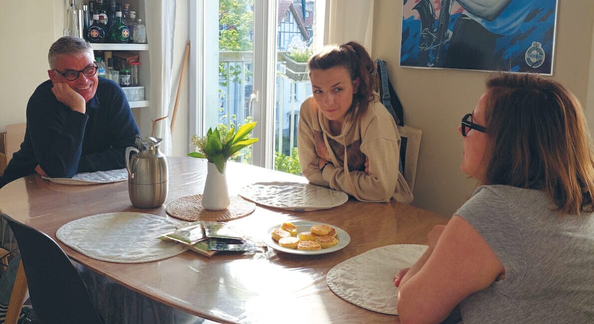 Tre personer sidder omkring et spisebord i et soloplyst rum. En vase med blomster og en tallerken med kager står på bordet. De ser ud til at være engageret i samtale. Et stort vindue og farverige kunstværker er synlige i baggrunden.