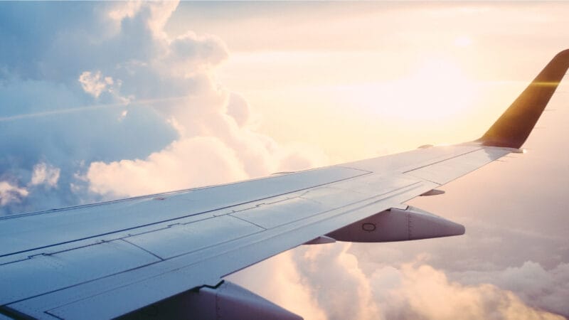 Udsigt fra et flyvindue, der viser en vinge mod en baggrund af luftige skyer og en gylden solnedgangshimmel. Som om den var lavet af en ekspert, kigger solen gennem skyerne og kaster et varmt skær på den fredfyldte scene.