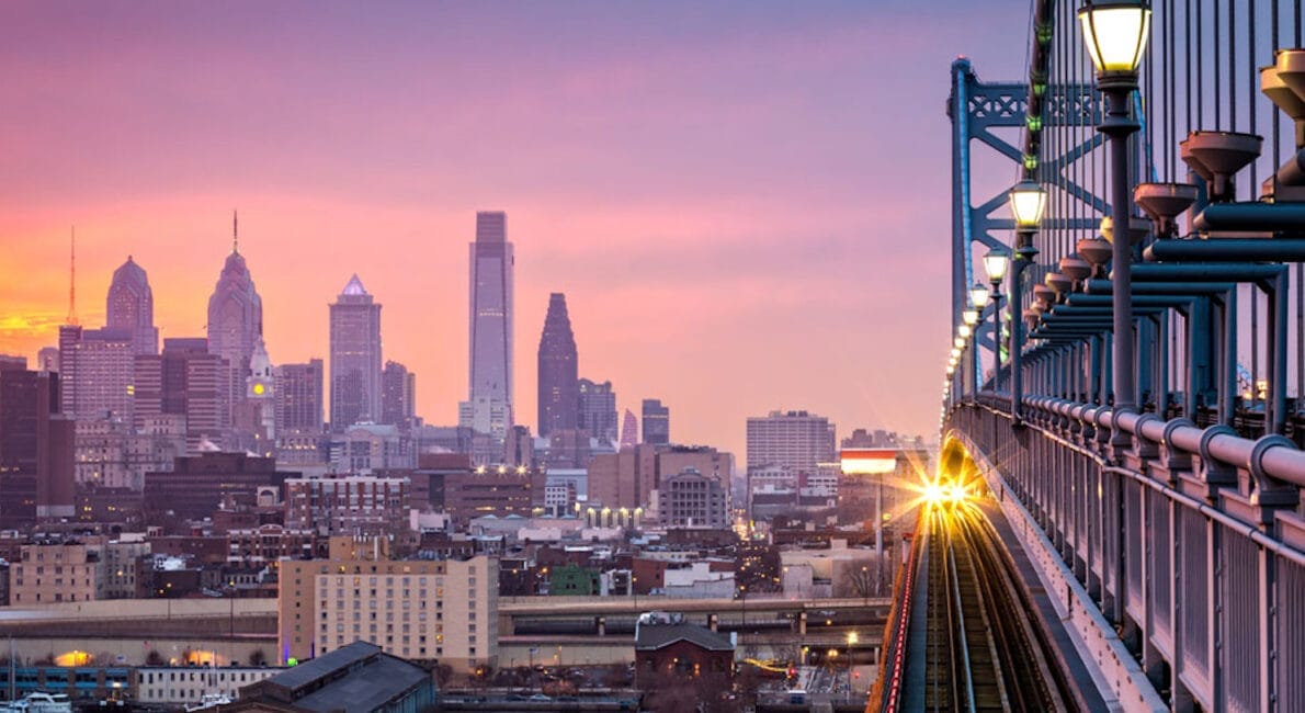 Et tog krydser en bro mod en pulserende byhorisont ved solnedgang, der minder om den betagende udsigt, som danskere verden over nyder. Bybilledet byder på tårnhøje skyskrabere mod en himmel malet i pink, lilla og orange nuancer, mens gadelygter smukt oplyser stien.