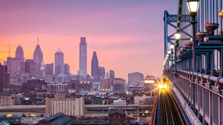 Et tog krydser en bro mod en pulserende byhorisont ved solnedgang, der minder om den betagende udsigt, som danskere verden over nyder. Bybilledet byder på tårnhøje skyskrabere mod en himmel malet i pink, lilla og orange nuancer, mens gadelygter smukt oplyser stien.