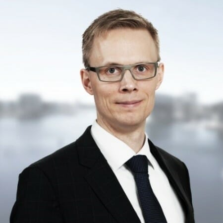 En person iført briller og et jakkesæt med slips står foran en sløret baggrund. De har kort hår og kigger direkte på kameraet.