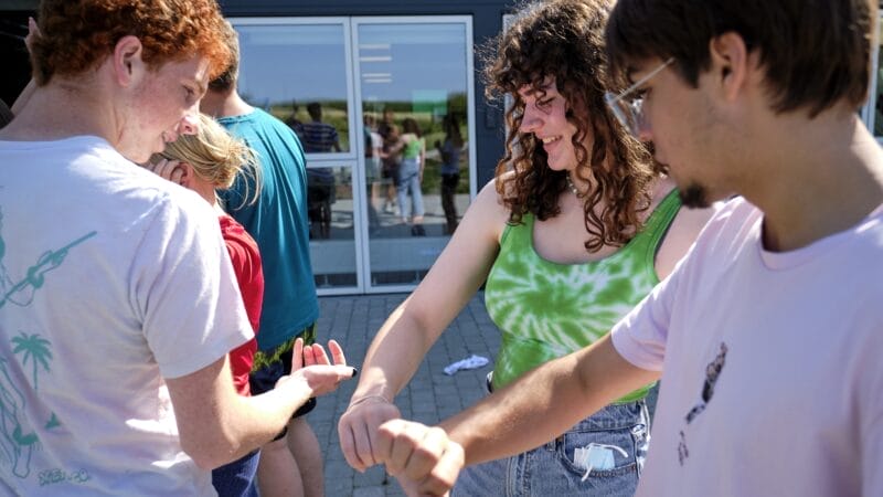 En gruppe mennesker står udendørs og smiler og rækker deres arme ud mod hinanden på en legende måde. De er afslappet klædt, og omgivelserne ser ud til at være i nærheden af en bygningsindgang med en glasdør.