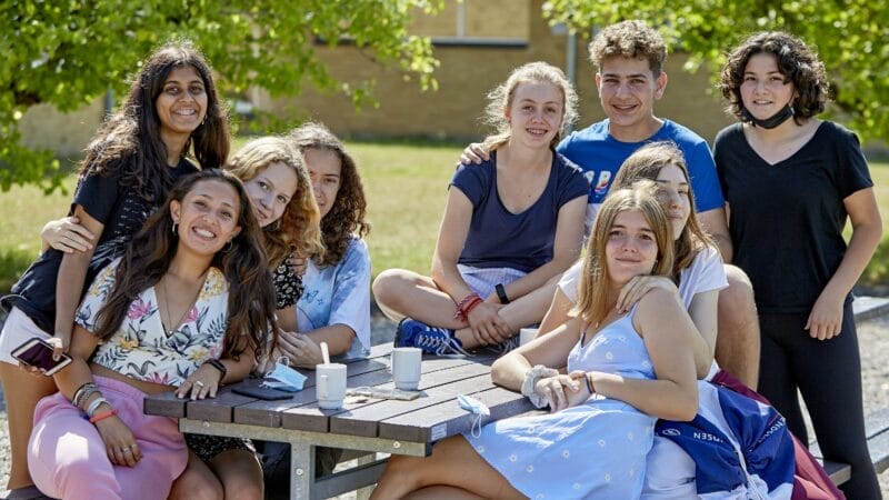 En gruppe på ni teenagere samledes afslappet omkring et picnicbord i træ udendørs og nød medlemsfordele af solskinsvejr og hinandens selskab. De er alle smilende og afslappede med grønne træer i kulissen og et par krus hvilende på bordet.