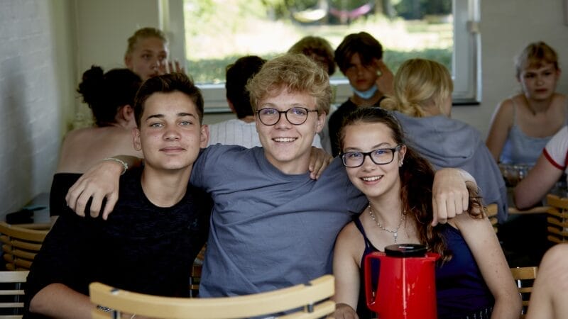 Tre personer fra den danske sommerskole sidder ved et bord og smiler til kameraet. To af dem har briller på, og en rød termokande er foran. Andre sidder i baggrunden, mens naturligt lys strømmer gennem et vindue.