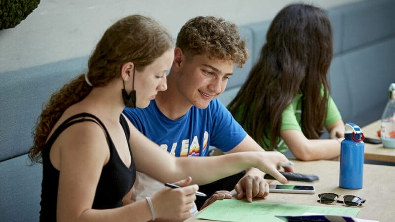 Tre personer ved et bord; to i forgrunden, den ene skriver på et grønt papir, den anden smiler. En ung kvinde i baggrunden kigger væk. Elementer på bordet inkluderer solbriller, en blå vandflaske og en telefon.