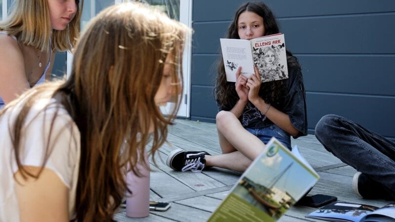 A group of young people sitting on a wooden deck, reading various books and comics. One girl holds a comic titled Ellens Ark, while the others focus on different books. The background shows a grey wall and a glass door.