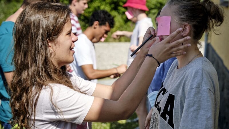 A group of young people outdoors. A girl places a sticky note on another girls forehead. Others in the background engage in similar activity. Theyre in casual clothes, with greenery behind them.