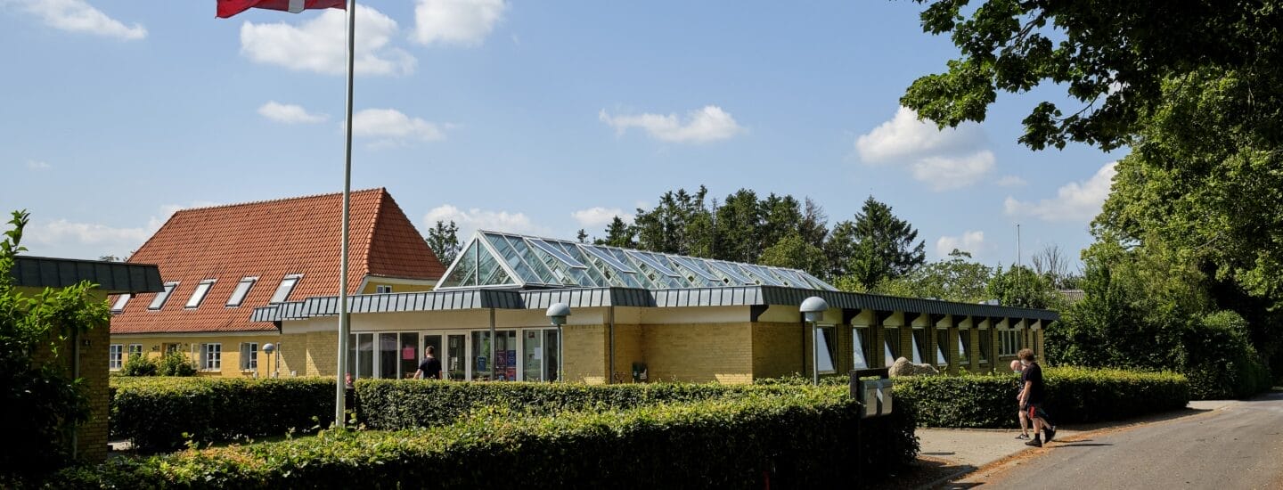 A building with a red-tiled roof and modern glass extension is surrounded by greenery under a clear blue sky. A Danish flag flies on a pole, and a person walks on a nearby road.