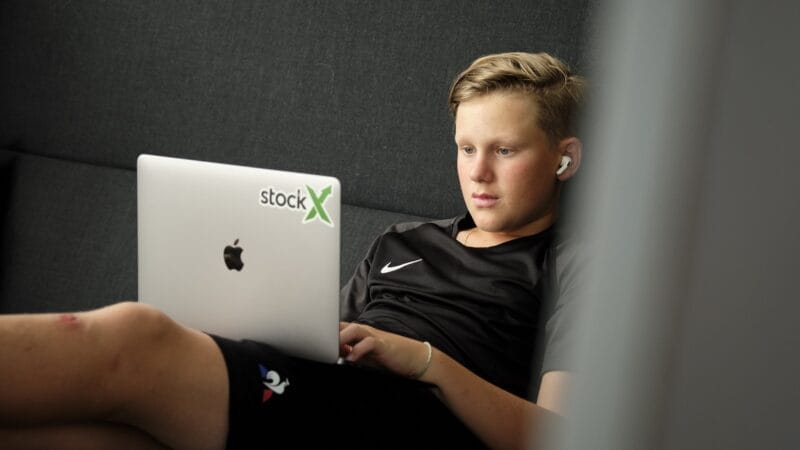 En person med kort hår ligger på en sofa ved hjælp af en bærbar computer, der er prydet med et "stockX"-klistermærke. Iført en sort Nike-skjorte og -shorts har de trådløse ørepropper i. Benene er draperet over sofaen, mens sollys filtrerer gennem vinduesrammen, der minder om den danske sommerskole-dage.