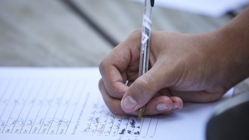A hand holding a pen, signing a document on a wooden surface. The paper contains multiple lines of text and signatures. The focus is on the hand and pen, with the background slightly blurred.