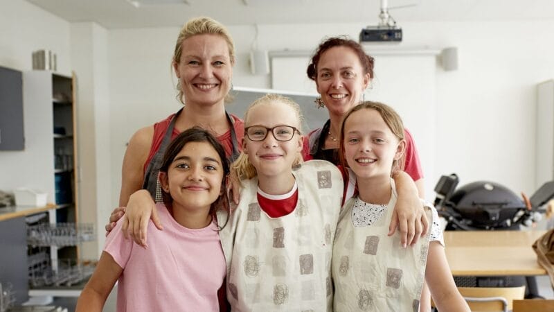 Two adults stand behind three smiling children in a classroom. The children wear aprons and appear happy, while the adults smile warmly. Tables and classroom equipment are visible in the background.