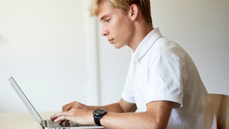 Ung mand med blond hår iført en hvid skjorte, sidder ved et bord og bruger en bærbar computer. Han ser fokuseret ud på skærmen med et ur på venstre håndled. Baggrunden er ude af fokus, hvilket giver et minimalistisk, fredfyldt miljø.