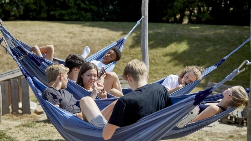 En gruppe på syv unge mennesker slapper af i to tilstødende blå hængekøjer i et græsklædt område. Nogle sidder op og taler og griner, mens andre læner sig tilbage og hviler sig. Træer og sollys er synligt i baggrunden.