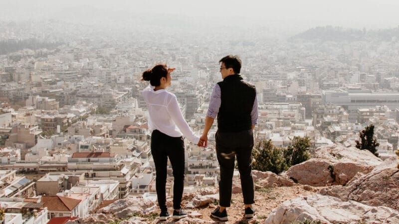 A couple stands on a rocky hill, holding hands and looking out over a densely built cityscape. The city sprawls into the distance under a hazy sky. The woman shields her eyes from the sun with her hand.