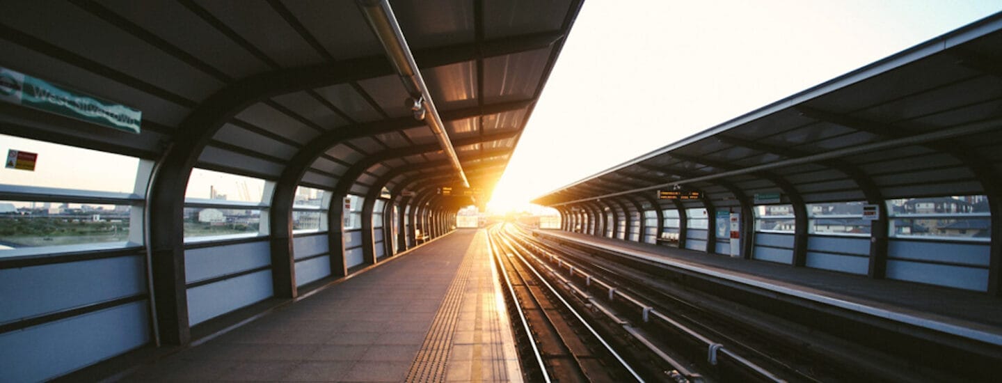 En moderne togstationsperron ved solnedgang, hvor den nedgående sol kaster lange skygger og et varmt skær på skinnerne og perronen. Stationen er tom, med buede tag med glaspaneler, der hvælver sig over sporene – en perfekt kulisse til at betragte livets mærkesager.