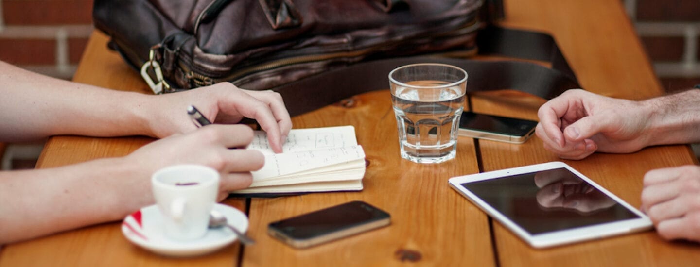 To personer sidder ved et træbord med en notesbog, pen, smartphone og tablet. Et glas vand og en kop kaffe er i nærheden. En lædertaske hviler på bordet. Rammerne foreslår et afslappet studiemøde eller studievejledningsmøde.