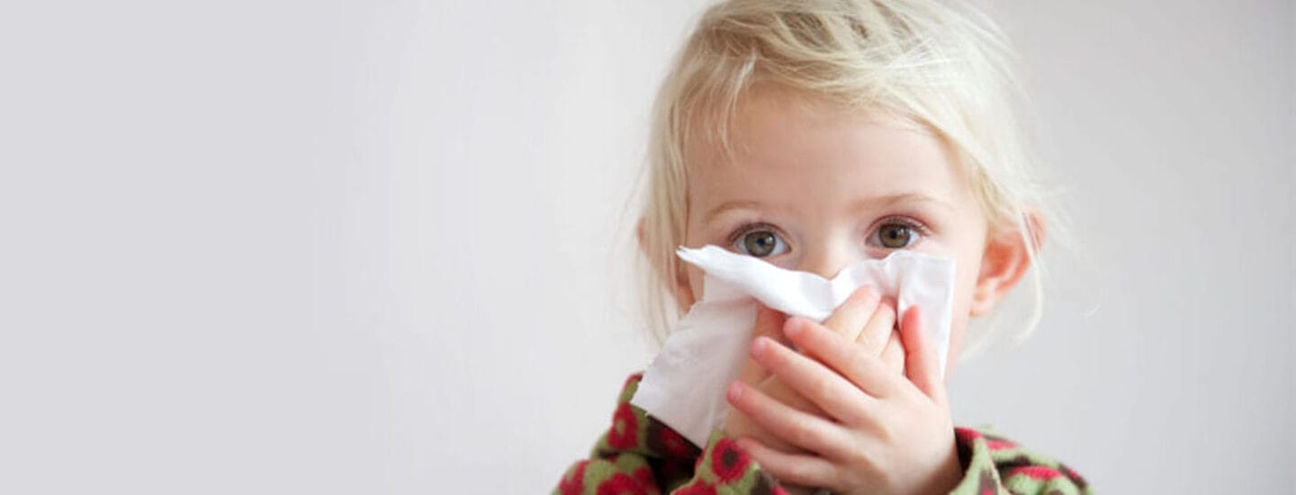 A young child with blonde hair uses a white tissue to wipe their nose. Theyre wearing a floral-patterned sweater, standing against a plain background, likely waiting for some advice from Hejdoktor.