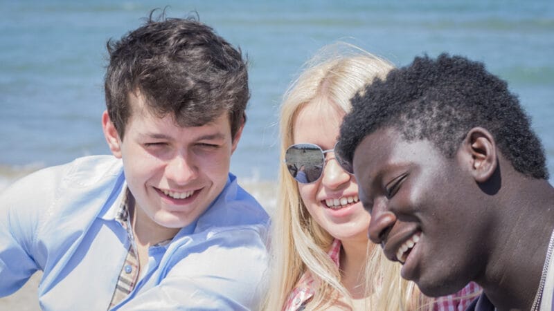 Three friends, looking like theyve just wrapped a lively student counseling session, are smiling and leaning together at a beach. One has dark curly hair, another has blonde locks with sunglasses, and the third boasts short, textured black hair. The ocean serenely stretches under a bright sky.