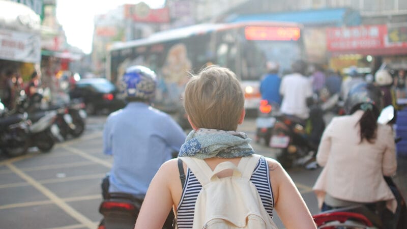 A person with short hair, wearing a striped top and a scarf, carries a white backpack. They navigate the bustling urban landscape, surrounded by motorcycles and a bus. Their presence blends seamlessly into the citys vibrant membership of daily commuters.