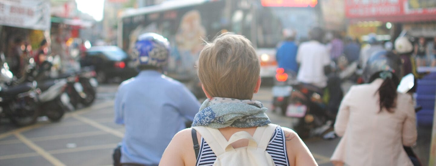A person with short hair, wearing a blue scarf and white backpack, stands amid a bustling network of motorbikes and a bus. The scene is vibrant with people in helmets and blurred store signs in the background.