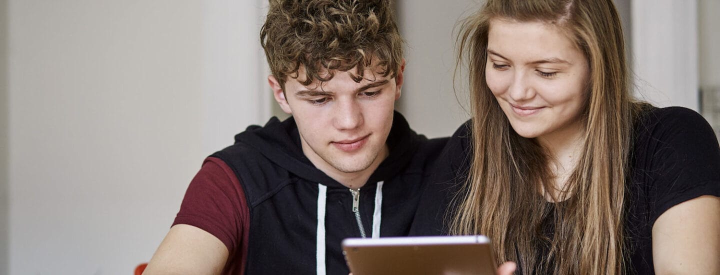 Two young people sit closely, engrossed in a tablet that explains school level Danish: how does it work? The person on the left wears a black hoodie, while the one on the right has long hair and a striped shirt. They are seated indoors with light-colored walls as their backdrop.