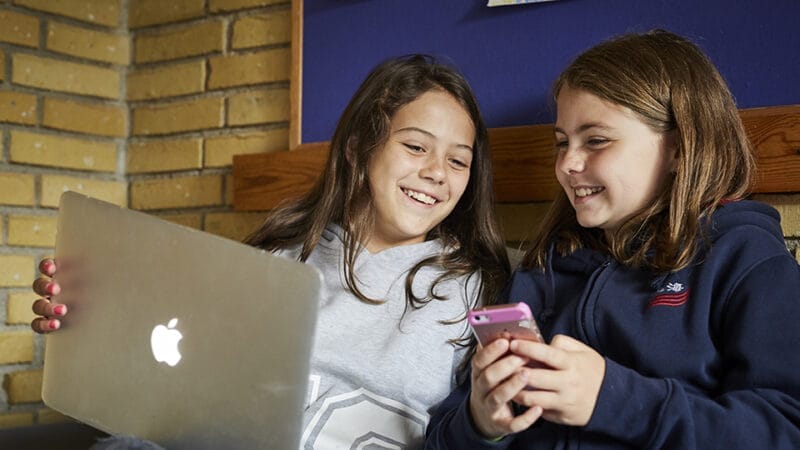 To piger sidder på en sofa og smiler, mens de deler noget på skærmen. Den ene holder en bærbar med et Apple-logo, og den anden tjekker sin telefon. They're likely exploring vejledning til skoledansk. En murstensvæg og træhylde fuldender de hyggelige rammer.