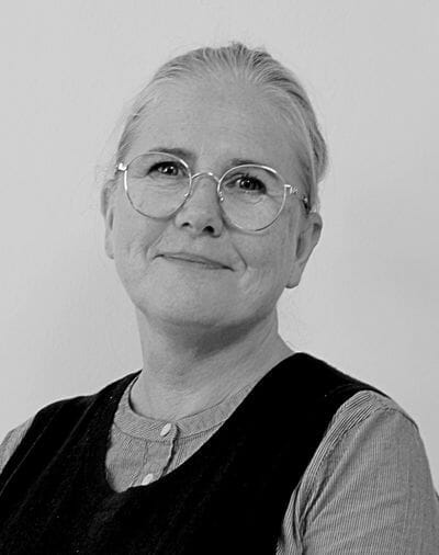 En kvinde med briller og et blidt smil kigger mod kameraet. Hun har lyst hår trukket tilbage, er iført en stribet top og en mørk vest og står på en almindelig baggrund. Billedet er i sort/hvid.