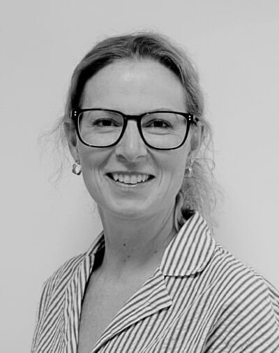 En person med langt hår smiler, bærer briller og en stribet skjorte på et sort/hvidt foto. Baggrunden er almindelig.