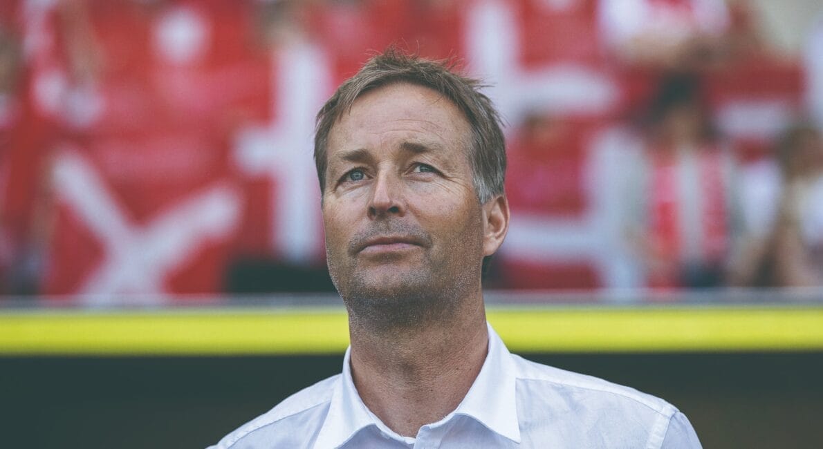 En mand i en hvid skjorte står foran slørede røde og hvide stadionsæder. Baggrunden har utydelige former og farver, der antyder livlige omgivelser. Hans udtryk er roligt og kontemplativt.