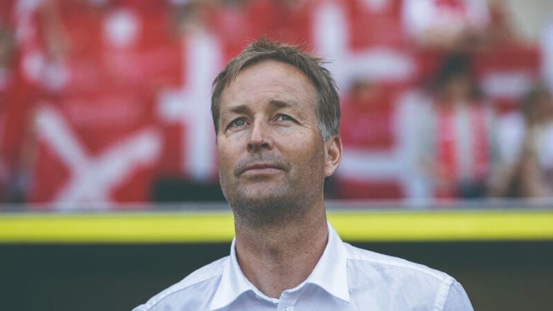 En mand i en hvid skjorte står foran slørede røde og hvide stadionsæder. Baggrunden har utydelige former og farver, der antyder livlige omgivelser. Hans udtryk er roligt og kontemplativt.