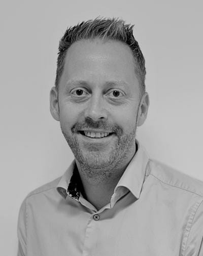 Black and white photo of a smiling person with short hair and a beard, wearing a light-colored collared shirt.