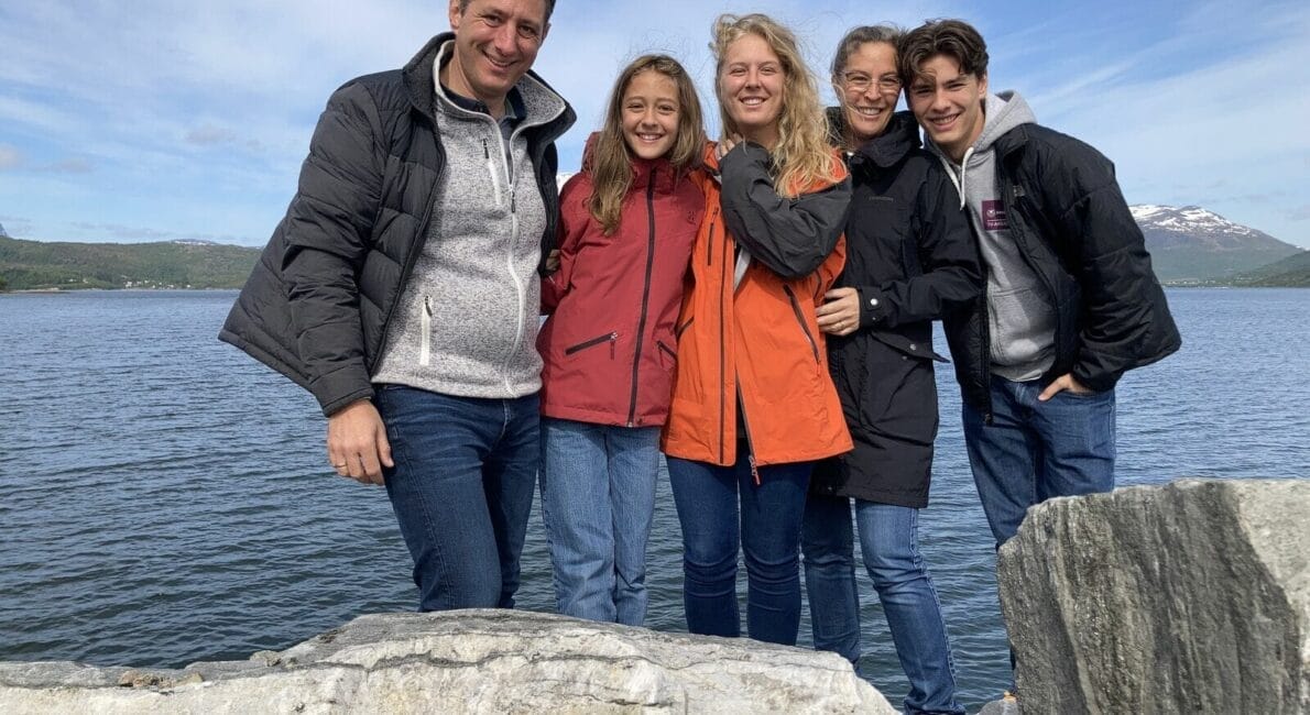 En gruppe på fem personer, to mænd og tre kvinder, smiler og poserer sammen udendørs. De står på klipper ved en sø, under en delvist overskyet himmel, med bjerge i baggrunden. De er klædt i afslappede jakker og jeans.