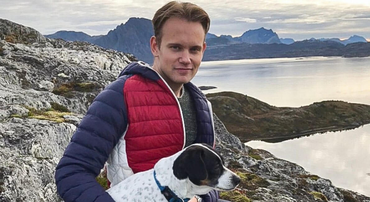 En person i en quiltet jakke holder en hvid og sort hund, mens han sidder på stenet terræn. I baggrunden er der en naturskøn udsigt over en sø omgivet af bjerge under en overskyet himmel.
