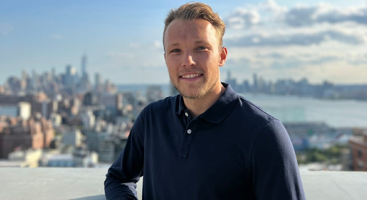 En person med kort hår og skæg smiler, mens han står på et tag med byens skyline og vand i baggrunden. De har en mørkeblå langærmet skjorte på. Himlen er delvist overskyet.