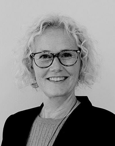 A black and white portrait of a person with short, curly hair and glasses, smiling at the camera. They are wearing a dark blazer over a textured sweater. The background is plain.