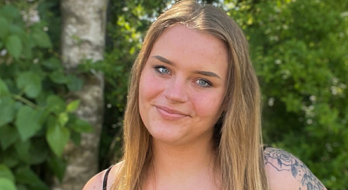 En person med langt hår og en tatovering på skulderen smiler til kameraet, mens han står udendørs. Frodigt grønt løv og sollys giver en lys, naturlig baggrund.