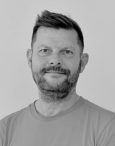 Et portræt i gråtoner af en mand med skæg og kort hår, iført en almindelig T-shirt, og kigger direkte på kameraet med et lille smil. Baggrunden er en enkel, lysegrå tone.