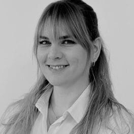 Sort og hvidt portræt af en person med langt, lyst hår og pandehår, smilende. De er iført en skjorte med krave og små øreringe. Baggrunden er almindelig.