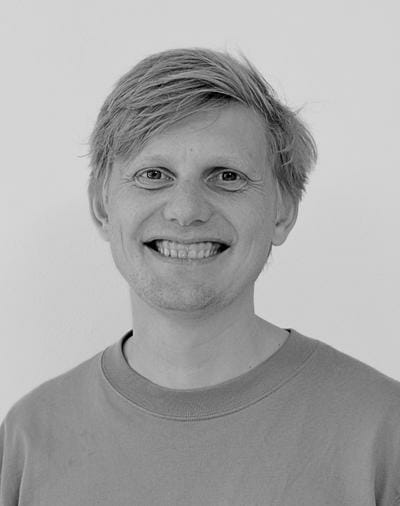 Sort/hvid foto af en smilende person med lyst hår, iført en almindelig skjorte, mod en enkel baggrund.