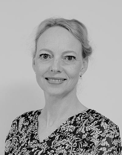 Black and white portrait of a smiling woman with light-colored hair tied back. She is wearing a patterned dress with floral designs and is looking directly at the camera. The background is a plain light color.