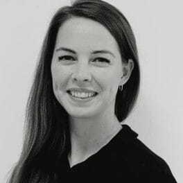 A black and white portrait of a person with long, straight hair, smiling and facing the camera. They are wearing a dark-colored top and small hoop earrings. The background is plain.
