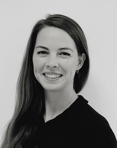 A black and white portrait of a person with long, straight hair, smiling and facing the camera. They are wearing a dark-colored top and small hoop earrings. The background is plain.