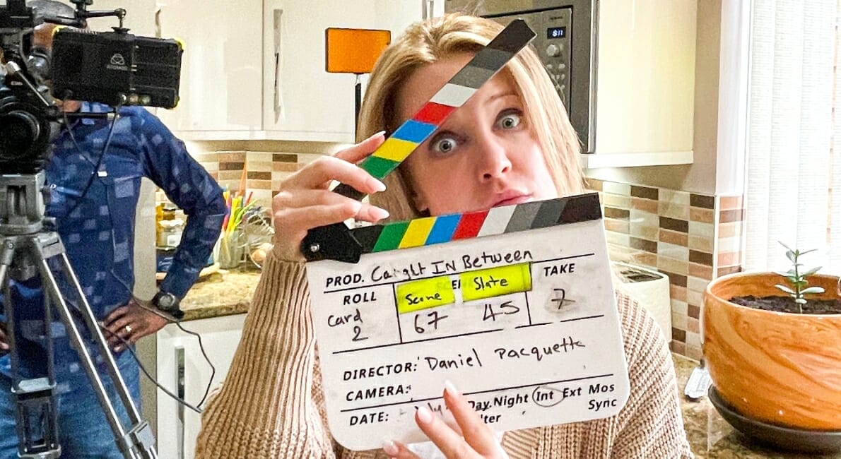 En kvinde i et køkken holder en filmklap med mærket "Caught In Between" og tavleoplysninger. Hun kigger legende gennem klappen. Der er et kamera og et besætningsmedlem til venstre og en potteplante til højre.
