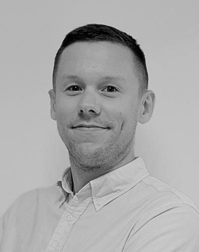 En person med kort hår smiler let og kigger mod kameraet. De er iført en lys skjorte med knapper. Baggrunden er ensartet og neutral, hvilket skaber et enkelt og professionelt udseende. Billedet er i sort/hvid.