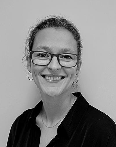 A person with glasses and hoop earrings smiles at the camera. They are wearing a dark shirt and a thin necklace. The background is plain and light-colored. The image is in black and white.