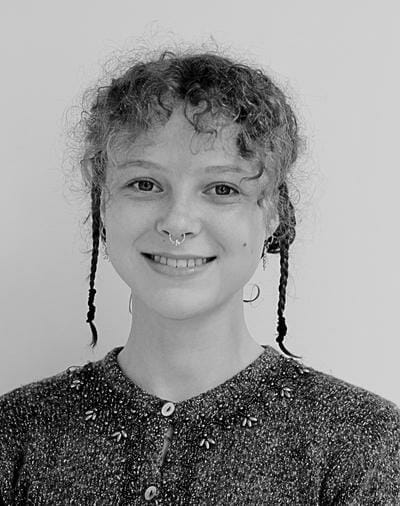 Young person with curly hair and braids smiles. They have a nose ring and wear a textured sweater with embroidered details. Black and white photo with a plain background.