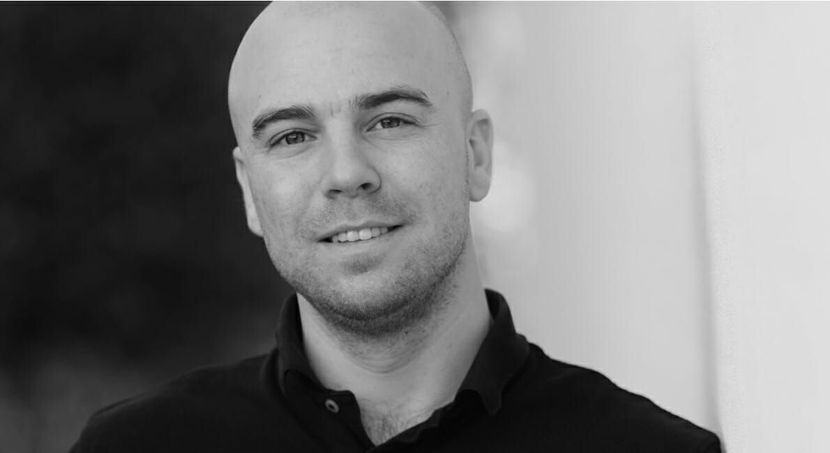 Et sort-hvidt billede af en skaldet mand med kort skæg, der smiler til kameraet. Han er iført en mørk skjorte med krave og står på en sløret baggrund.