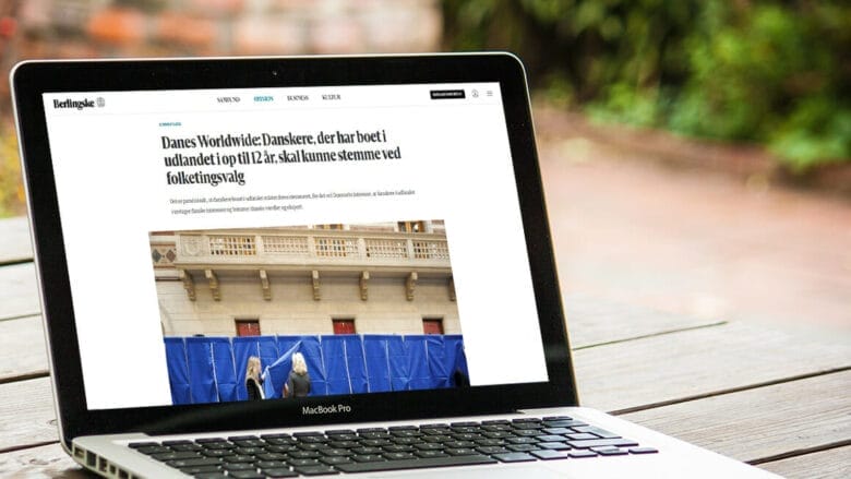 En bærbar, der viser en artikel på sin skærm. Overskriften nævner danskere, der har boet i udlandet i 12 år og stemt ved valg. Billedet på skærmen viser folk bag stemmebokse. Den bærbare computer står på et træbord udendørs.