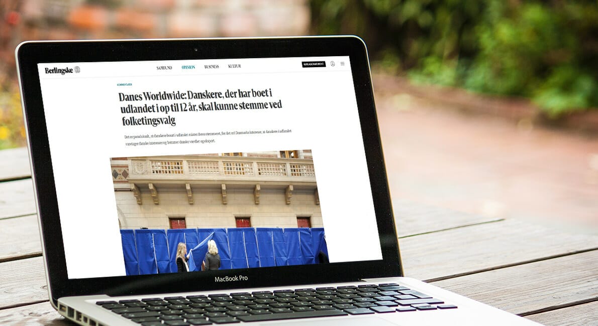 En bærbar, der viser en artikel på sin skærm. Overskriften nævner danskere, der har boet i udlandet i 12 år og stemt ved valg. Billedet på skærmen viser folk bag stemmebokse. Den bærbare computer står på et træbord udendørs.