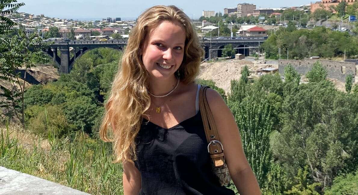 En person med langt, bølget blond hår smiler og står udendørs foran en naturskøn udsigt. Bag dem er frodige grønne træer, en bro og et bybillede under en klar blå himmel. De har en sort top på og holder en taske.